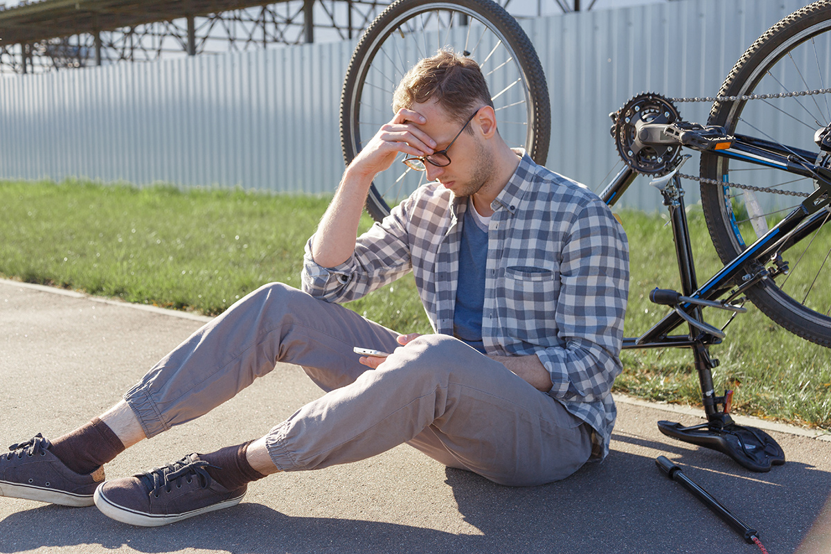 A puzzled man searches the internet for the cause of a bicycle malfunction while sitting on the road. The bike is turned over against the background.